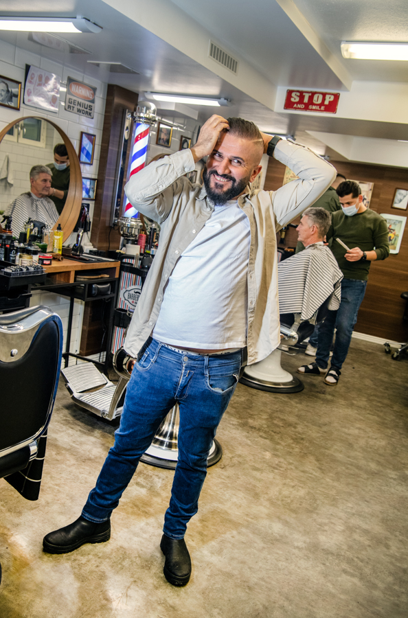 Ivan står i sin barberar- och frisörsalong. Han håller armarna ovanför huvudet då han precis dragit en kam igenom håret. I bakgrunden står en frisör och klipper en man. 