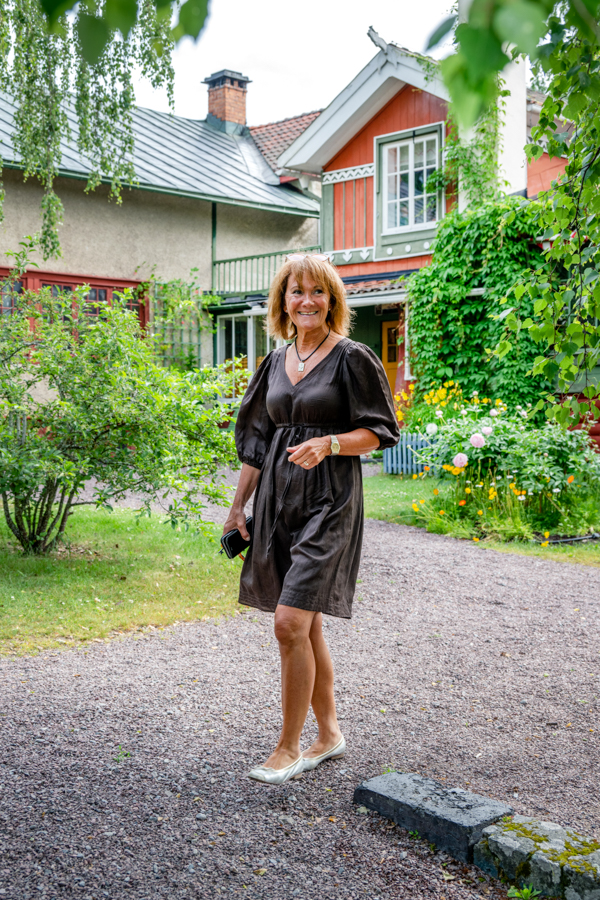 Chia går på grusgången i trädgården vid Carl Larsson-gården. Hon har en mörkgrå knälång klänning på sig. I bakgrunden syns klätterväxter på den faluröda husfasaden, blomsterrabatter och buskar.