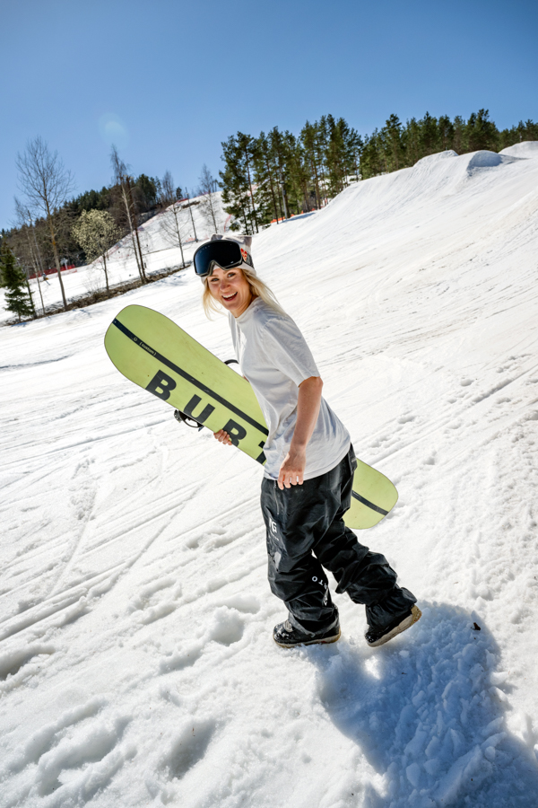 Ylfa går i Källviksbacken men sin snowboardbräda under armen. Snön ligger vit och i bakgrunden syns de stora hoppen i backen.