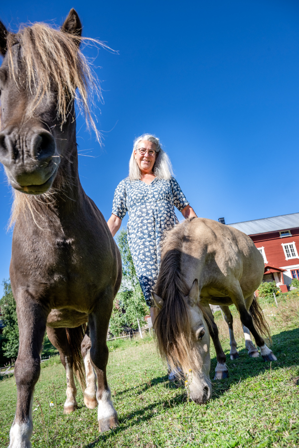 Maria står i hästhagen, klappar på två hästar och tittar in i kameran. Den svarta hästen står nära kameran och tittar även den in i kameran. Den bruna hästen står precis bredvid Maria och äter gräs. 