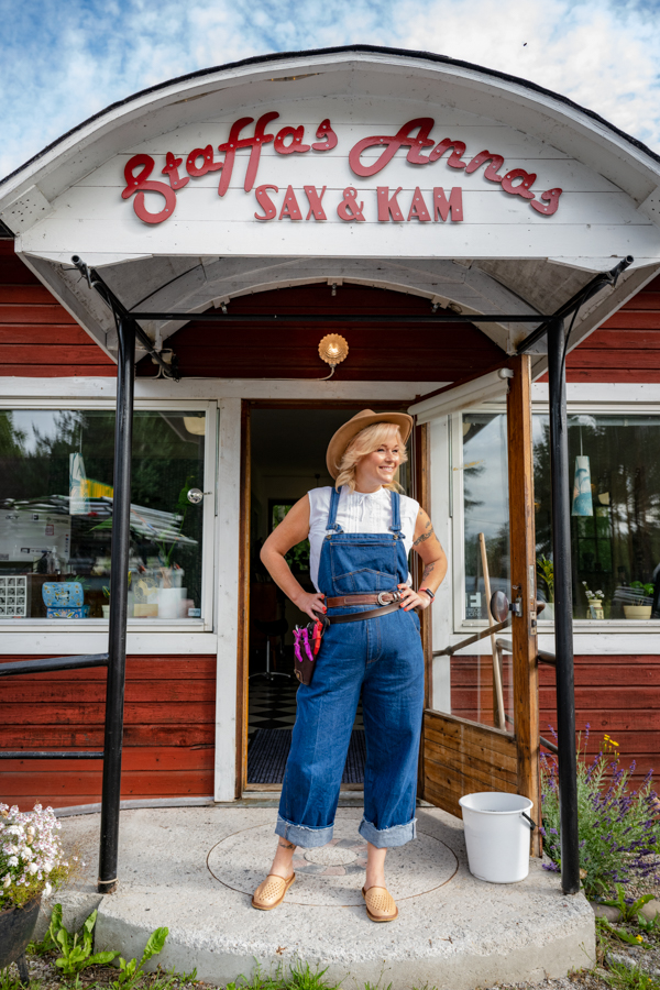 Anna står på farstukvisten till sin frisersalong som ligger i en Faluröd stuga. Ovanför farstukvisten sitter en skylt där det står "Staffas Annas Sax & Kam".
