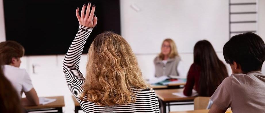 Flicka ses bakifrån räcka upp handen i ett klassrum