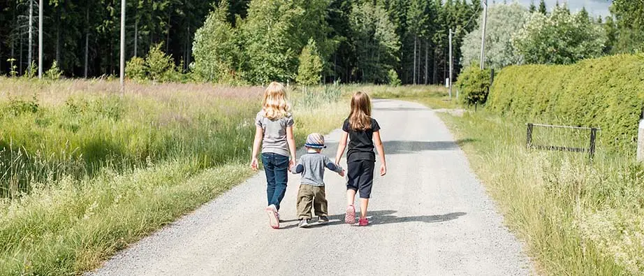 Tre barn som håller varandra i händerna och går på liten väg på landet.
