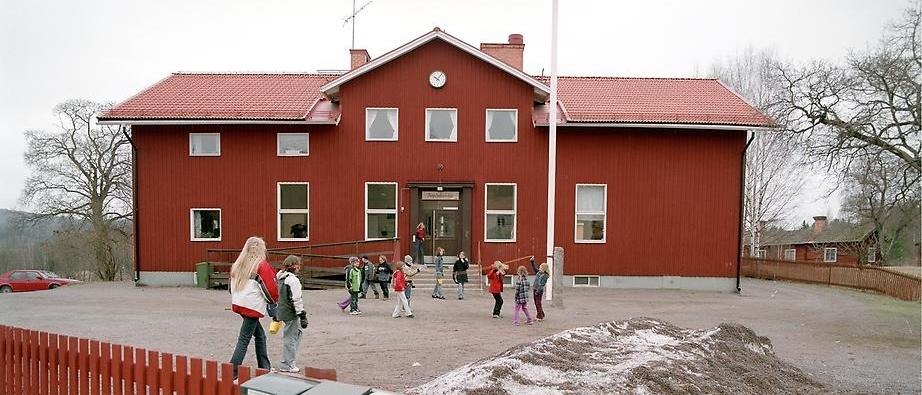 Bild över Aspebodaskolan. Skolan är faluröd och på gårdsplanen, framför skolan, leker barn. 