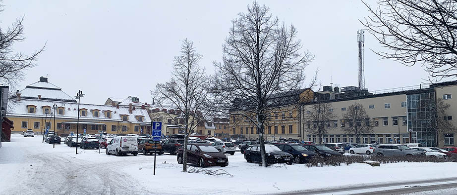 Många bilar på en stor parkering. Snö och vinter. I bakgrunden syns äldre byggnader i gul och vit färg.