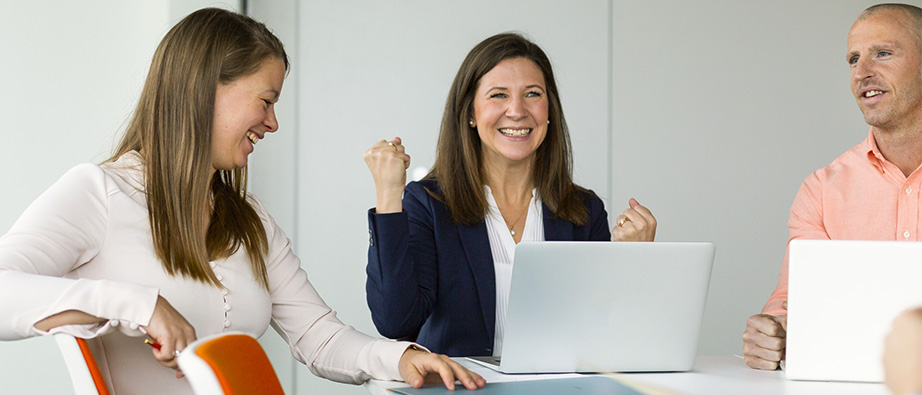 Två kvinnliga kollegor och en manlig vid arbetsbord med datorer. En kvinna gör tecken men handen som visar att något lyckats.