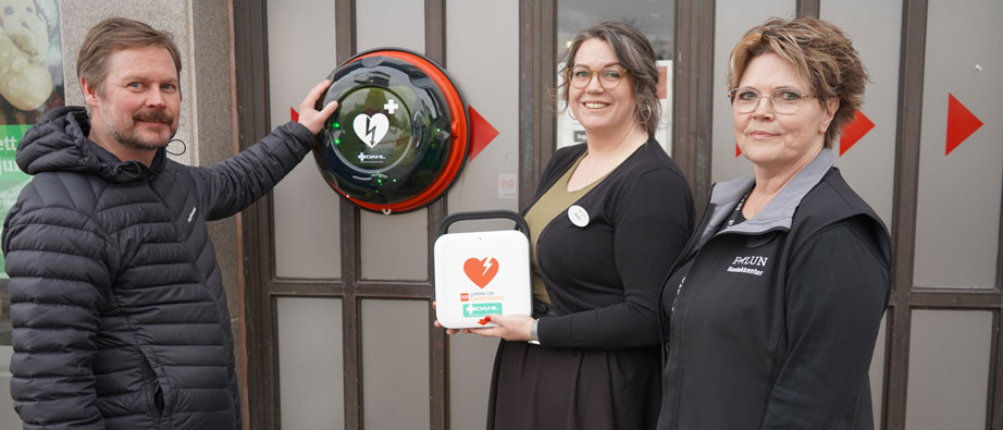 Tre personer som visar den hjärtstartare som är fäst på väggen utanför kontaktcenter och den hjärtstartare man kan låna