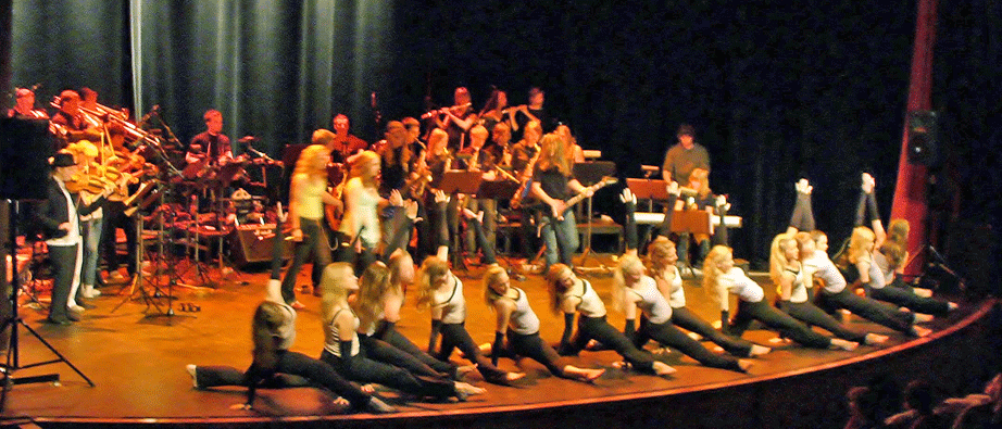Många personer som spelar, dansar och sjunger på en scen i gulaktigt ljus